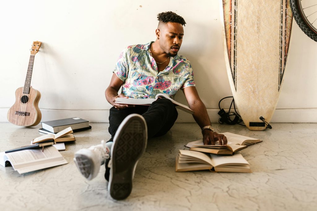 A young man sitting on the floor reading books, surrounded by a guitar and surfboard, embracing a creative and scholarly lifestyle.